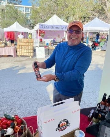Happy customer in a blue long-sleeve shirt and orange cap holding a bottle of Carlos Consuming Fire hot sauce at an outdoor market.