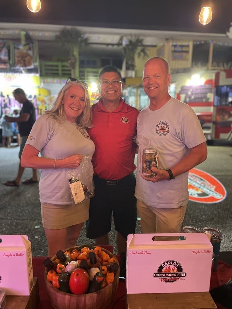 Carlos in a red polo shirt smiling with two happy customers holding Pepper Salada jars and hot sauce at an evening outdoor festival.