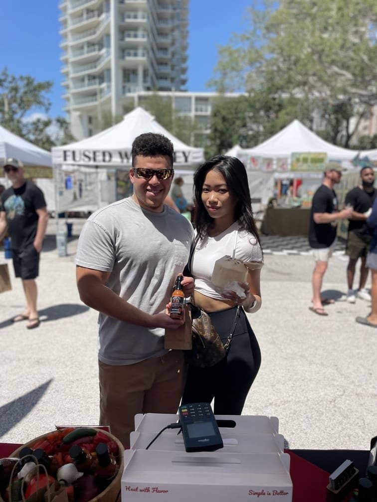 A happy young couple posing with a bottle of Carlos Consuming Fire Mango Habanero hot sauce at a sunny outdoor artisan market.