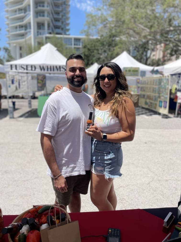 A happy couple smiling and posing with a bottle of Carlos Consuming Fire Hot Sauce at an outdoor community market.
