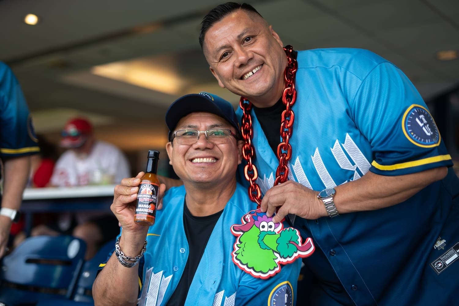 Carlos, owner of Carlos Consuming Fire, smiling with a fan in a blue Phillies jersey while holding a bottle of Mango Habanero hot sauce at a baseball game.