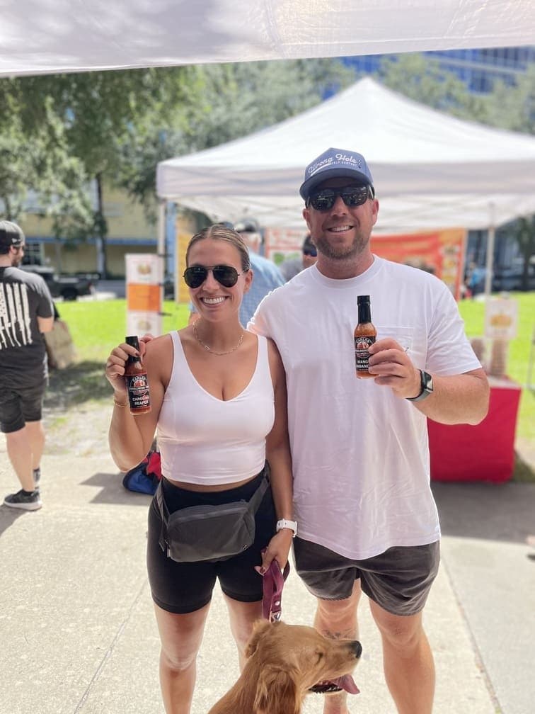 Happy couple holding two different bottles of Carlos Consuming Fire hot sauce while posing with their dog at an outdoor event.