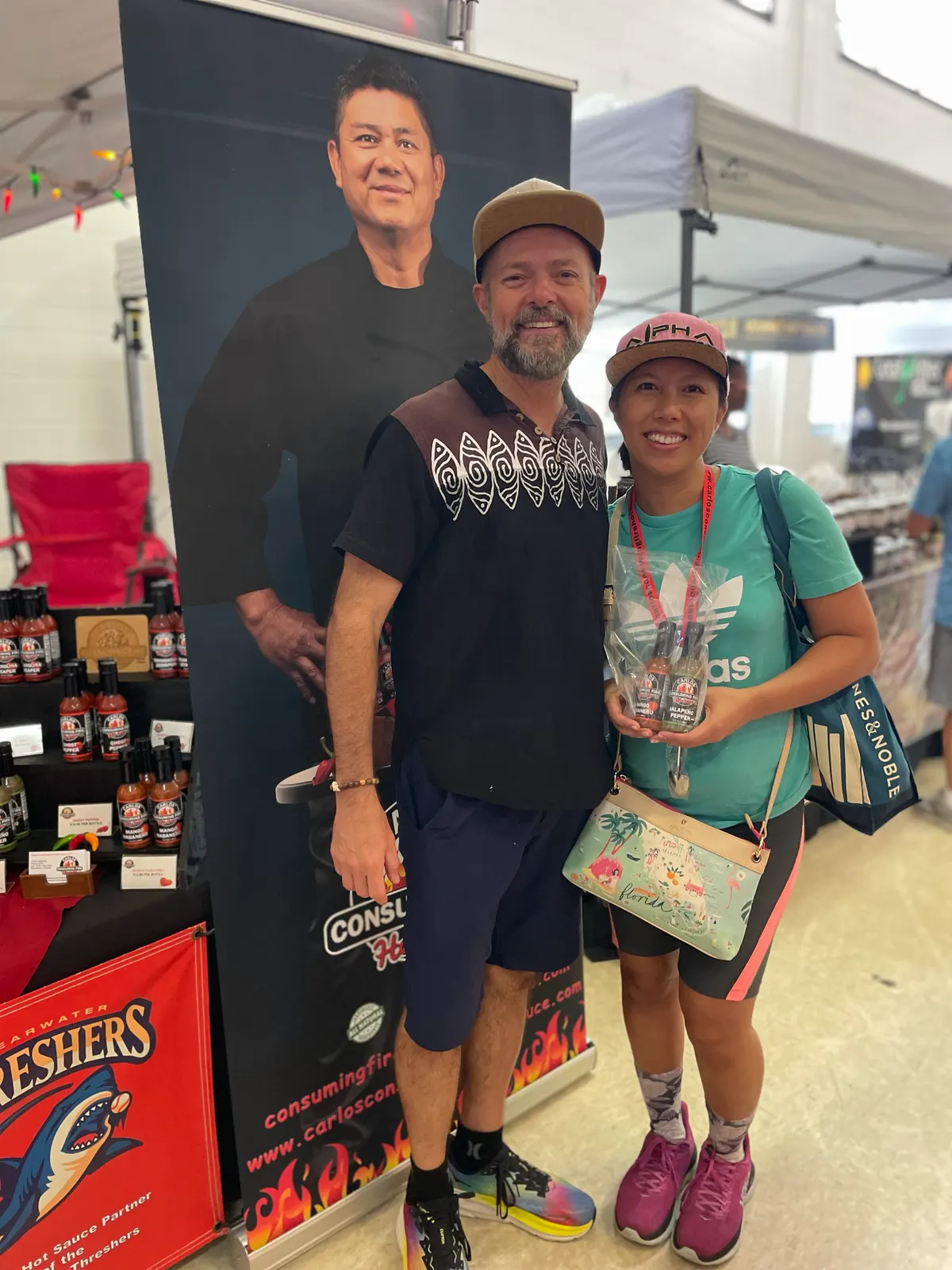 Portrait of a smiling couple posing with a wrapped gift pack of mini sauce bottles beside the Carlos roll-up banner and Threshers signage at an indoor event booth.