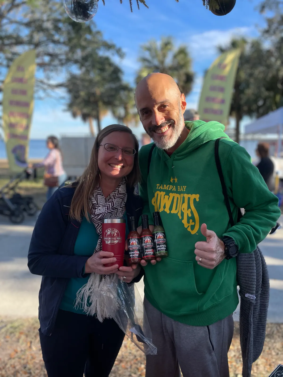 Portrait of a happy couple at an outdoor waterfront market holding a branded Carlos tumbler and a gift pack of three small sauce bottles, palm trees and tents behind; man gives a thumbs up.