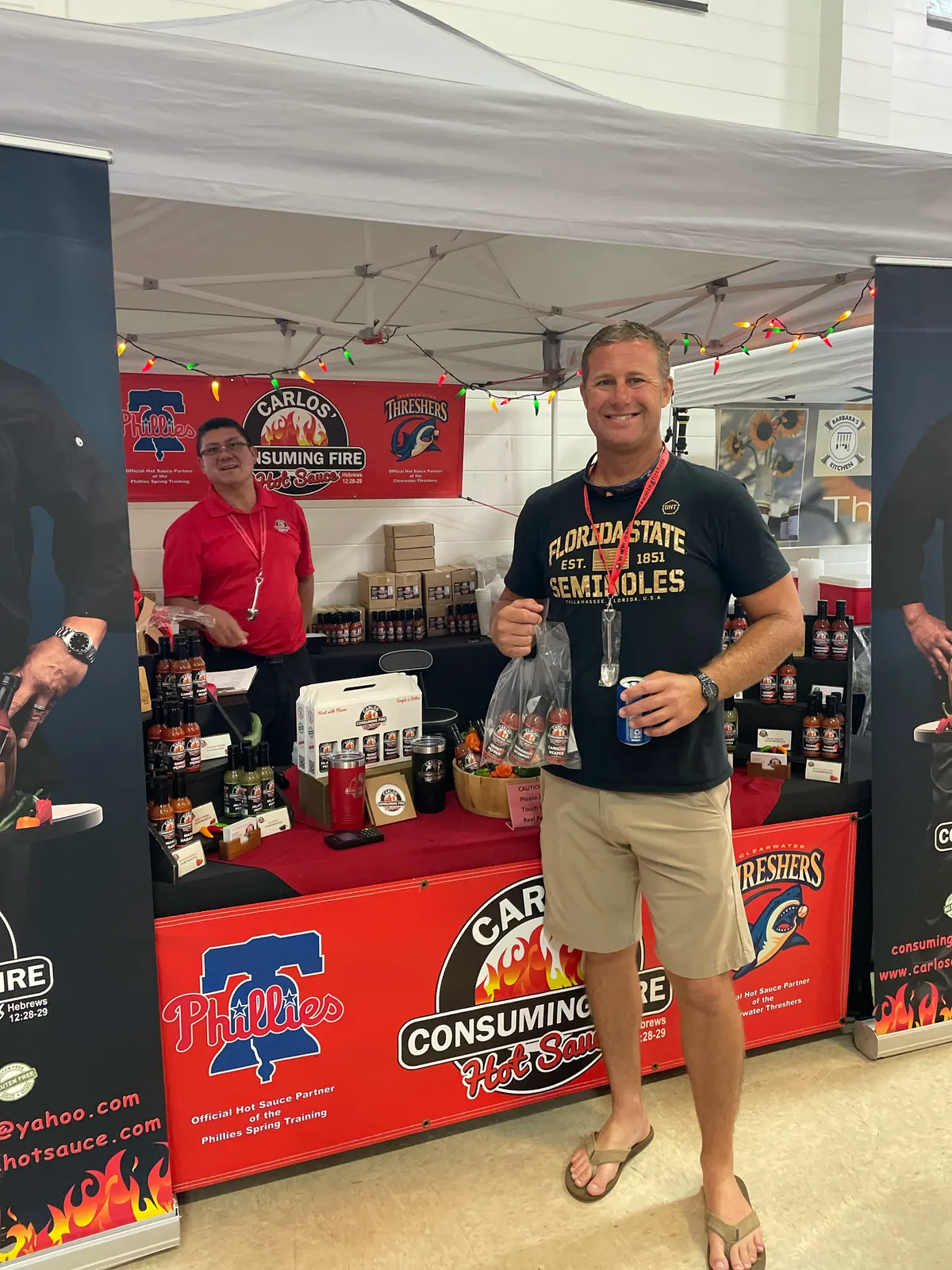 Portrait of a smiling customer in a Florida State shirt holding a bagged gift basket of sauces in front of the Carlos booth at an indoor market, with Phillies and Threshers partner banners and product tables visible.