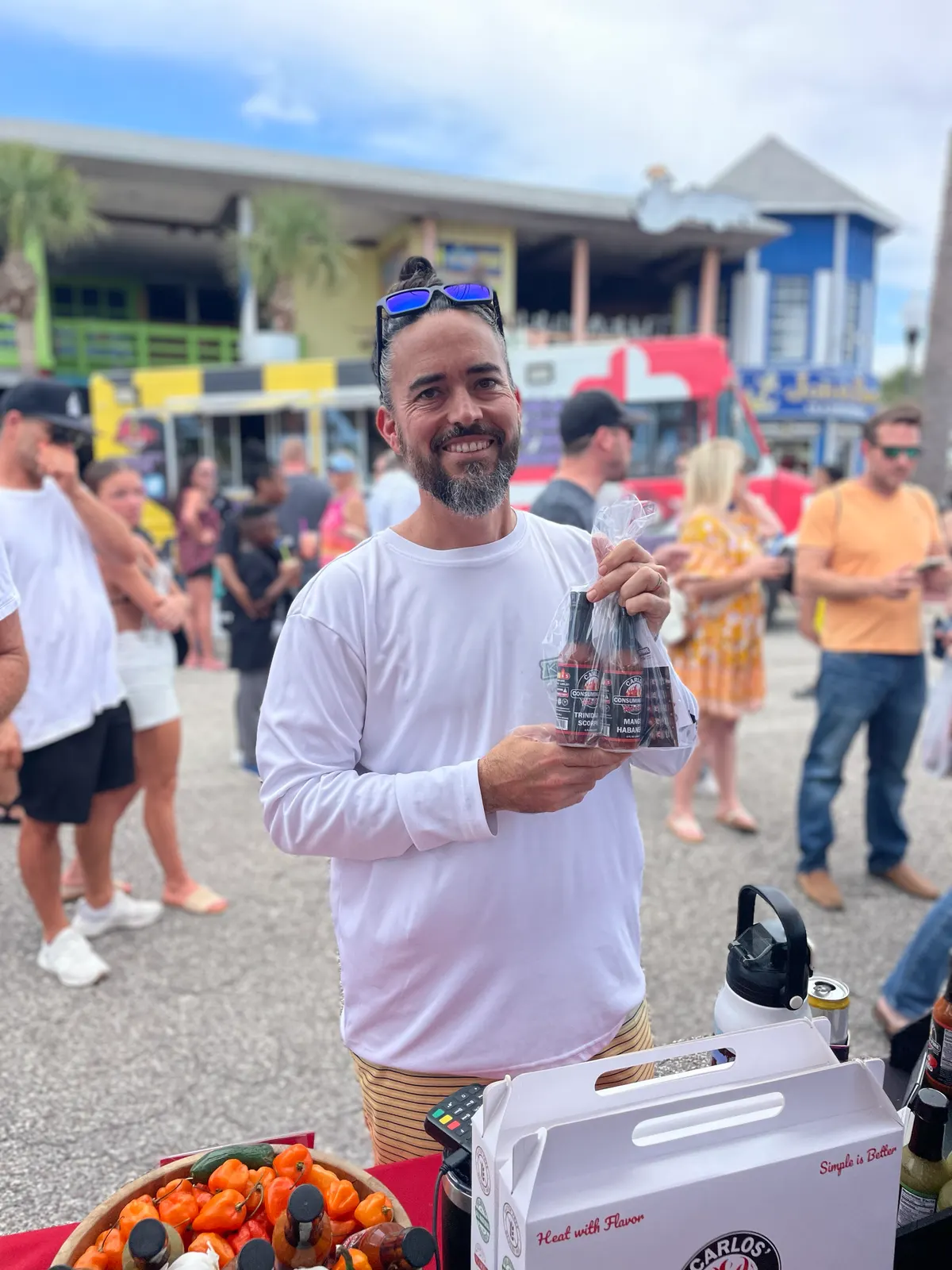 Portrait of a bearded customer in white holding a wrapped multi-bottle gift pack at the Carlos festival booth, branded white carry box and pepper basket in the foreground, lively boardwalk crowd behind.