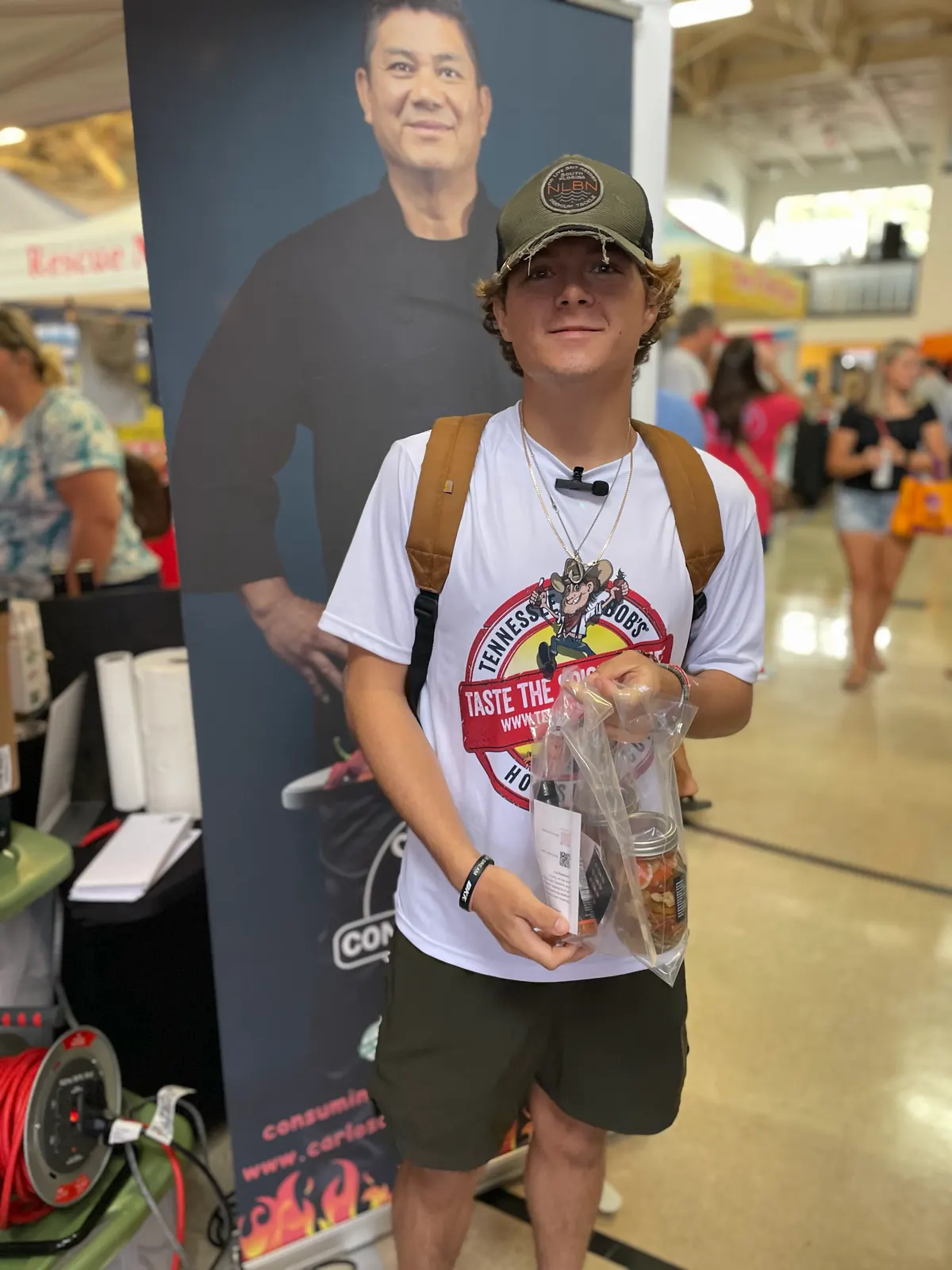 Portrait of a teenage customer in a cap holding a bagged jar/product in front of the tall Carlos' Consuming Fire roll-up banner (Carlos pictured) inside a busy market hall.