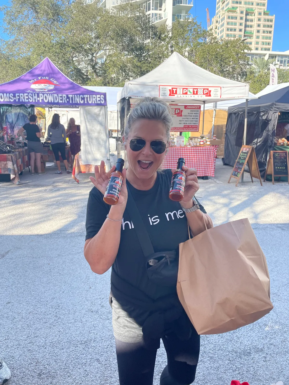 Portrait candid of a smiling customer in sunglasses at an outdoor farmers market holding up two red-label Carlos' bottles (variants unreadable) with vendor tents and city buildings behind; authentic customer-love shot.