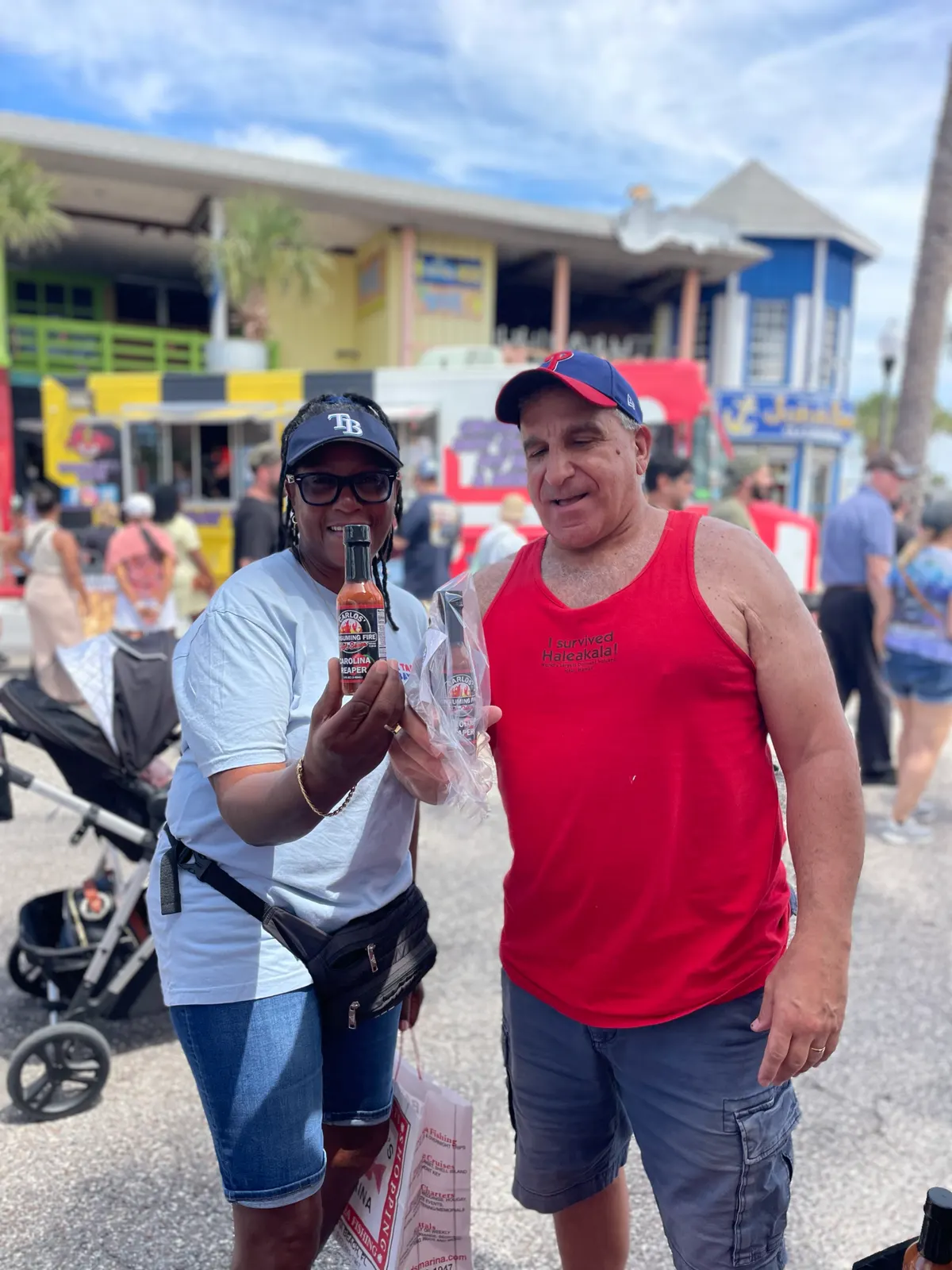 Portrait of two festival-goers (woman in Tampa Bay cap, man in red tank top) holding up a red-label sauce bottle and a bagged purchase at a colorful daytime boardwalk festival with food trucks behind.