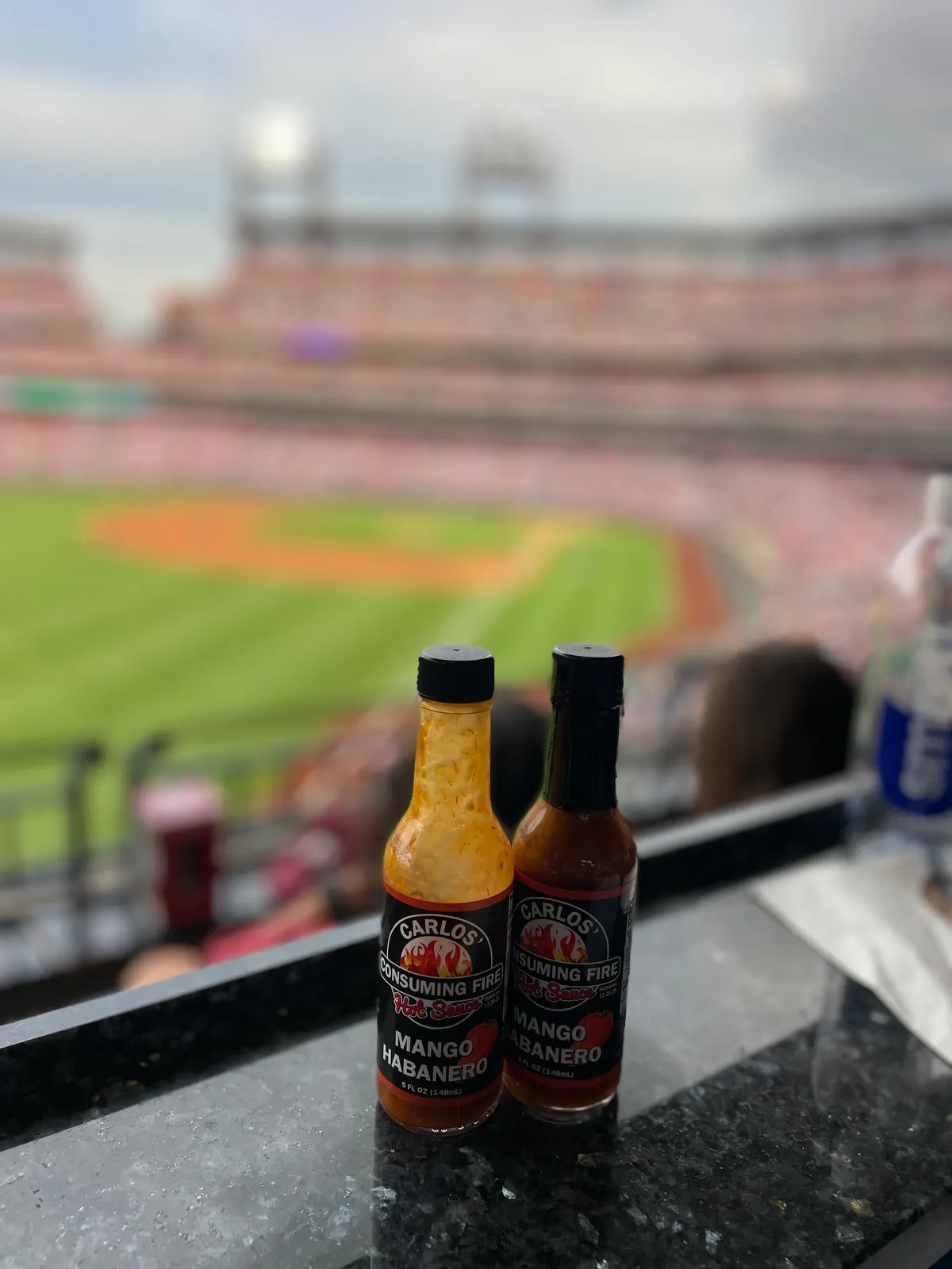 Hero-worthy portrait shot of two Carlos' Consuming Fire Mango Habanero bottles standing on a granite suite ledge with the ballpark field softly blurred behind.